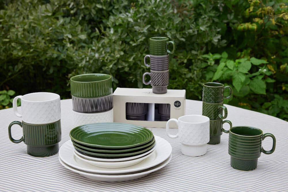 Ein Set aus grünem und weißem Keramikgeschirr, Schalen und Sagaform GmbH Coffee & More Teetassen (2-tlg.) steht auf einer gestreiften Outdoor-Tischdecke mit Blattpflanzen im Hintergrund; einige Tassen und Schalen sind gestapelt, daneben steht eine Coffee & More Box.