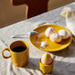 Ein Set gelber Sagaform Coffee & More Kaffeebecher (2-teilig, Sagaform GmbH), ein Teller mit dekorierten Eiern und Salz sowie ein Ei in einem gelben Eierbecher stehen auf einer beigen Tischdecke, das Sonnenlicht wirft sanfte Schatten – eine gemütliche Szene.