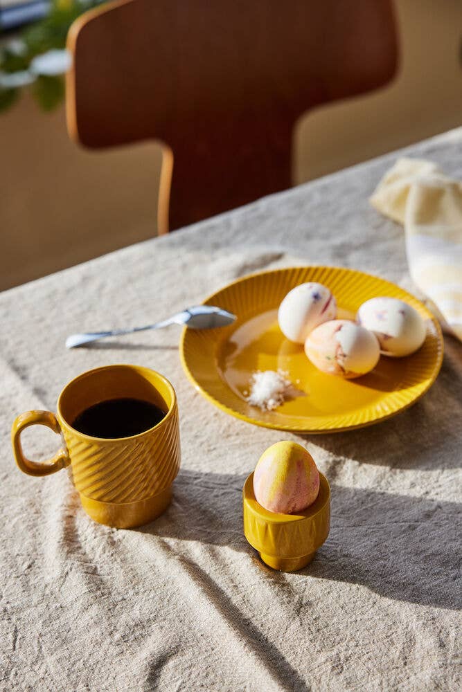 Ein Set gelber Sagaform Coffee & More Kaffeebecher (2-teilig, Sagaform GmbH), ein Teller mit dekorierten Eiern und Salz sowie ein Ei in einem gelben Eierbecher stehen auf einer beigen Tischdecke, das Sonnenlicht wirft sanfte Schatten – eine gemütliche Szene.