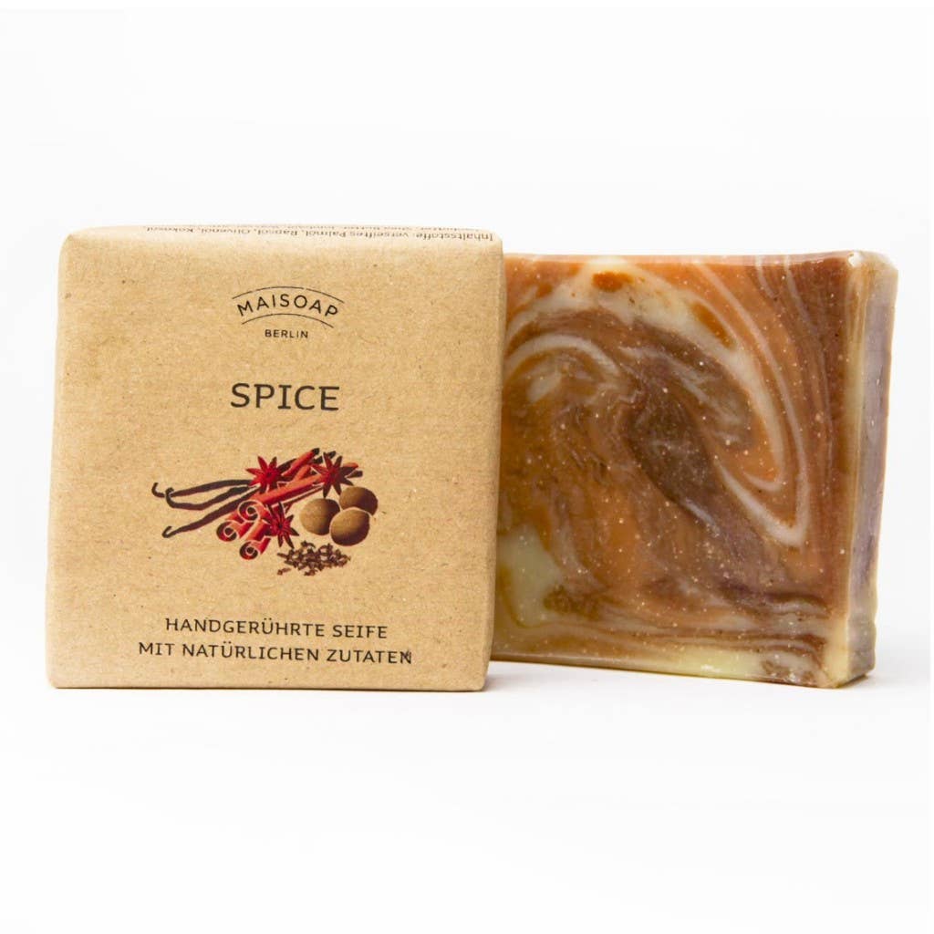 Eine quadratische Tafel marmorierter brauner und cremefarbener Gewürzseife von maisoap berlin steht neben ihrer hellbraunen Papierverpackung, die mit Gewürzillustrationen, deutschem Text über natürliche Inhaltsstoffe und einem Hauch von Nelke und Zimt verziert ist.