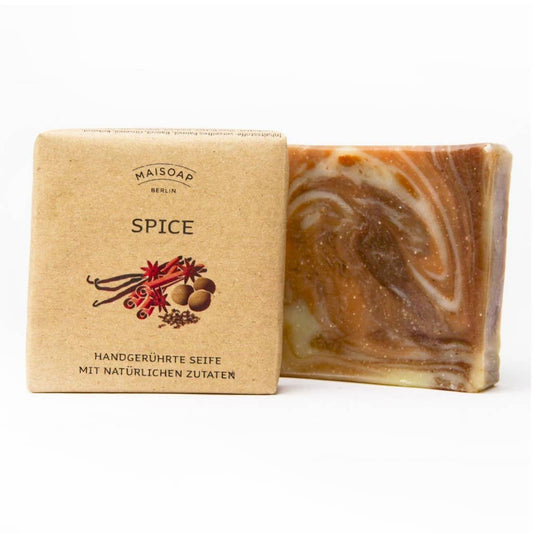 Eine quadratische Tafel marmorierter brauner und cremefarbener Gewürzseife von maisoap berlin steht neben ihrer hellbraunen Papierverpackung, die mit Gewürzillustrationen, deutschem Text über natürliche Inhaltsstoffe und einem Hauch von Nelke und Zimt verziert ist.