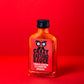 Eine 100-ml-Glasflasche der Crazy Bastard Sauce - Trinidad Scorpion & Clementine scharfe Soße von CBS Foods GmbH ruht auf einer roten Fläche mit einem roten Etikett mit fettgedrucktem schwarz-weißem Text und einer Illustration von großen Augen.