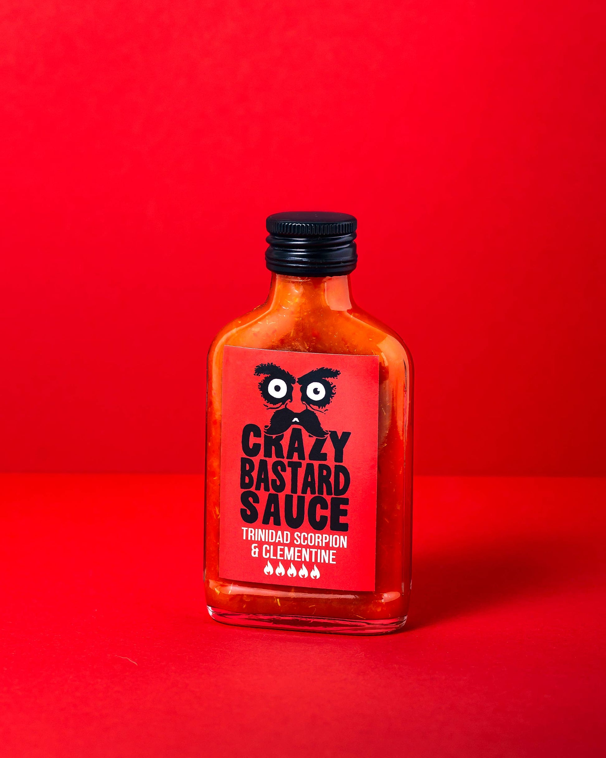 Eine 100-ml-Glasflasche der CBS Foods GmbH (Crazy Bastard Sauce) - Trinidad Scorpion & Clementine scharfe Soße steht vor einem leuchtend roten Hintergrund, ihr Etikett zeigt fette schwarz-weiße Schrift mit großen Augen über dem Markennamen.