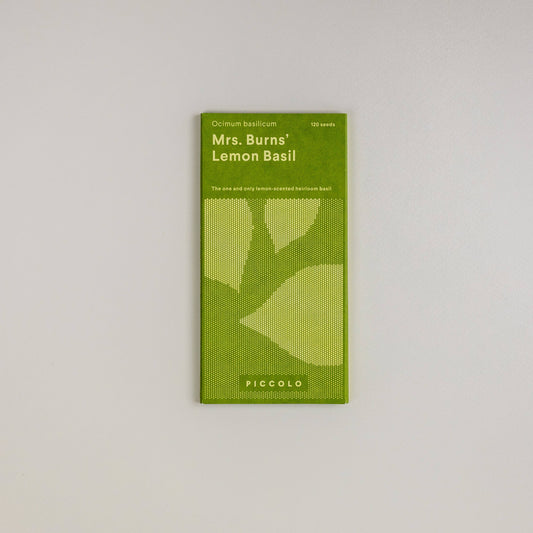 Eine grüne Packung Piccolo Seeds – Basilikum Mrs. Burns Zitrone liegt auf einem schlichten weißen Hintergrund und zeigt Blattillustrationen und Text, die den Status der Pflanze als Erbstück und den erfrischenden Zitronenduft hervorheben.