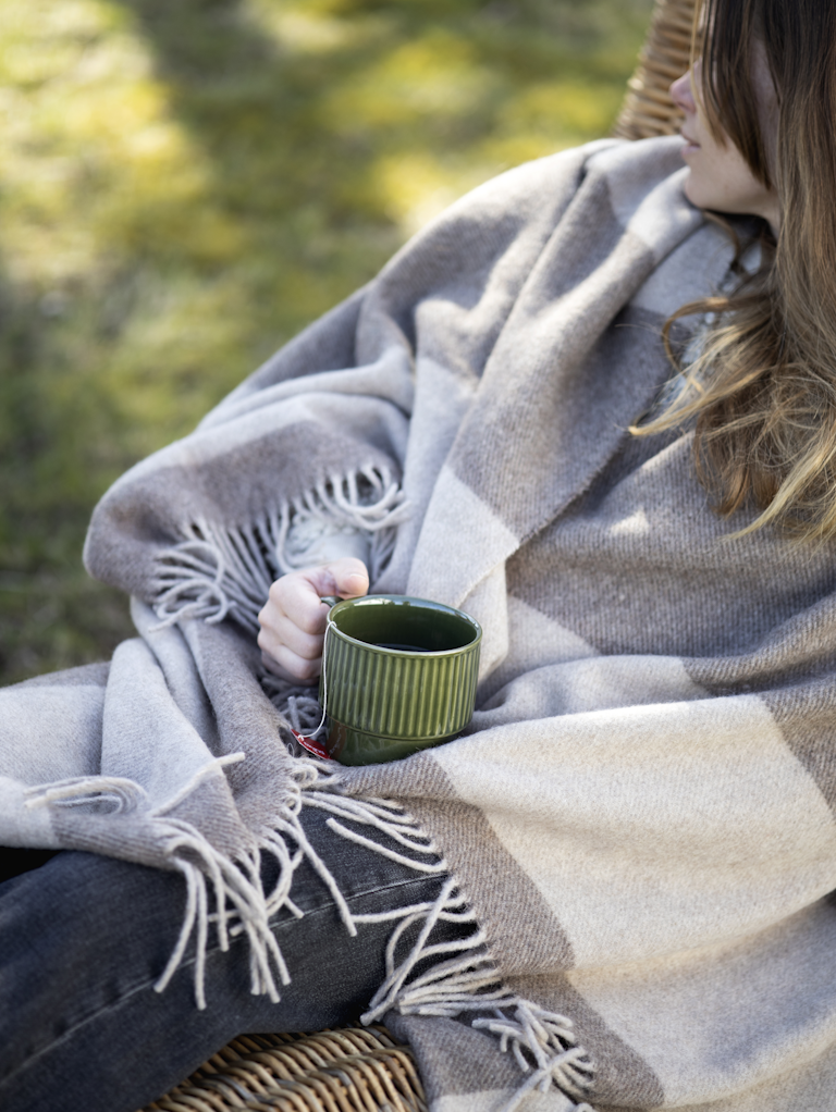 Eingehüllt in eine grau-beige karierte Decke sitzt jemand in einem Korbsessel im Freien und hält den Sagaform Coffee & More Kaffeebecher Grün, 2-tlg von Sagaform GmbH in der Hand - ein ruhiger, gemütlicher Moment vor einer sonnenbeschienenen Graskulisse.