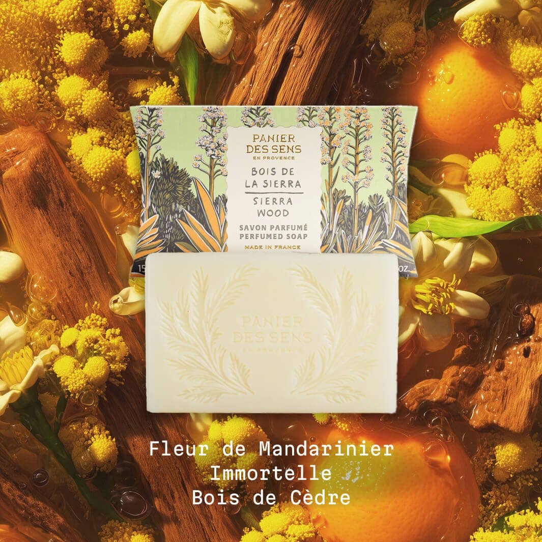 Ein Riegel Panier Des Sens Duftseife | Sierraholz 150 g ruht vor seiner illustrierten Schachtel, umgeben von Orangen, gelben Blüten und Holz. Der Text darunter lautet: Holziger Duft.