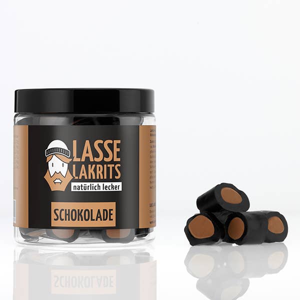 Ein durchsichtiges LASSE LAKRITS - natürlich lecker - Dose Lakritz Schokolade Glas von LASSE LAKRITS enthält Lakritzbonbons mit Schokoladenkern, die auf einer weißen Fläche neben dem Glas präsentiert werden.