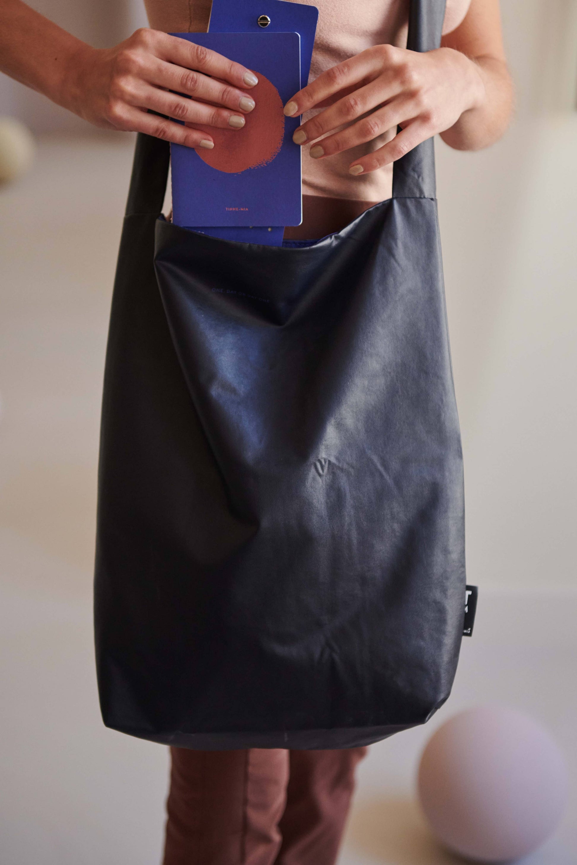 Eine Person hält eine dunkle Tasche und legt eine Tinne Mia Aufgabenliste in True Blue hinein. Sichtbar sind nur Oberkörper, Arme und ein Teil der Beine vor einem sanft verschwommenen Hintergrund.