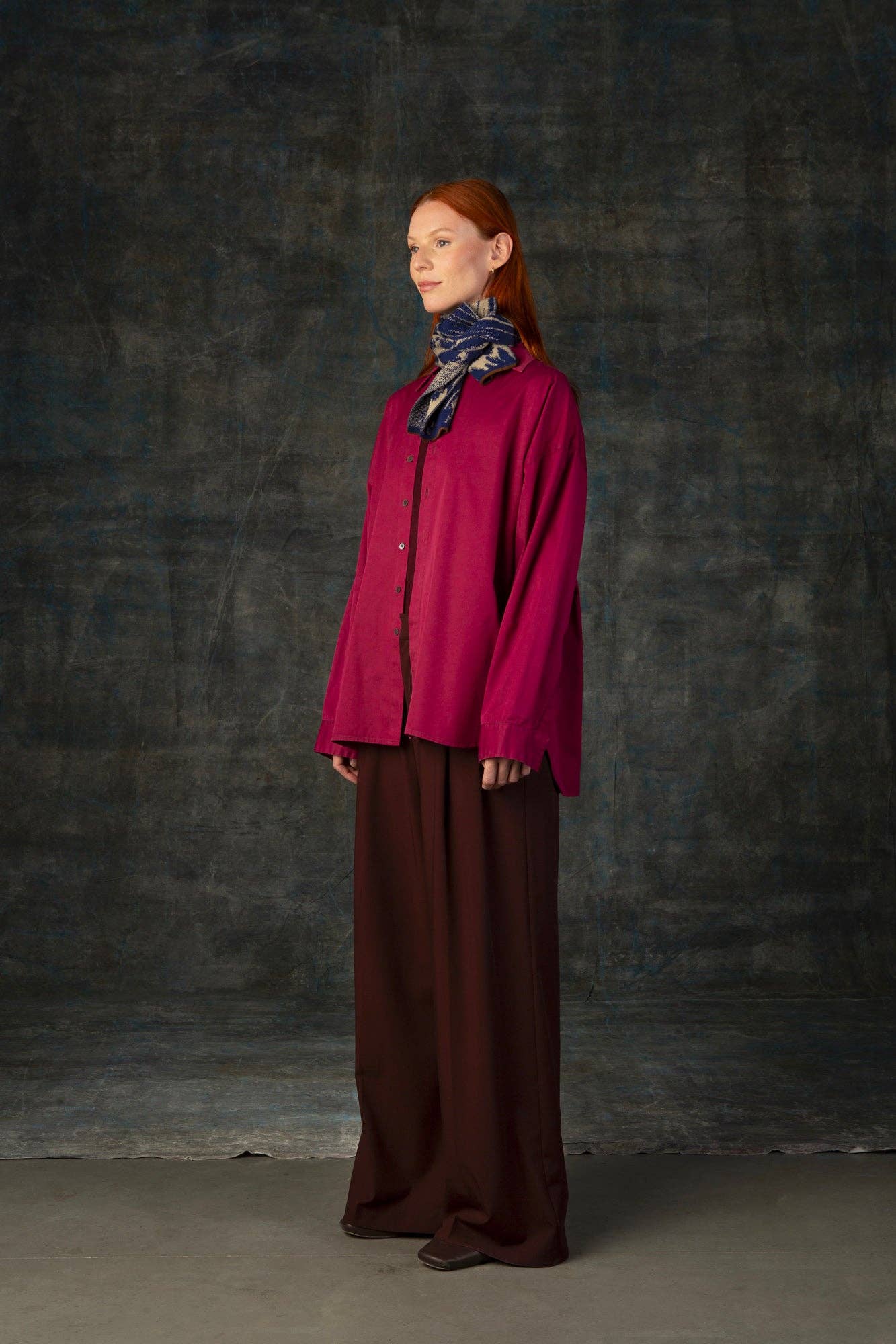 Eine Frau mit langen roten Haaren steht vor einem dunkel strukturierten Hintergrund. Sie trägt ein lockeres magentafarbenes Hemd, eine weitbeinige braune Hose, dunkle Schuhe und den Wolvis Schal aus Merinowolle - Aurata Indigo Mini um den Hals.