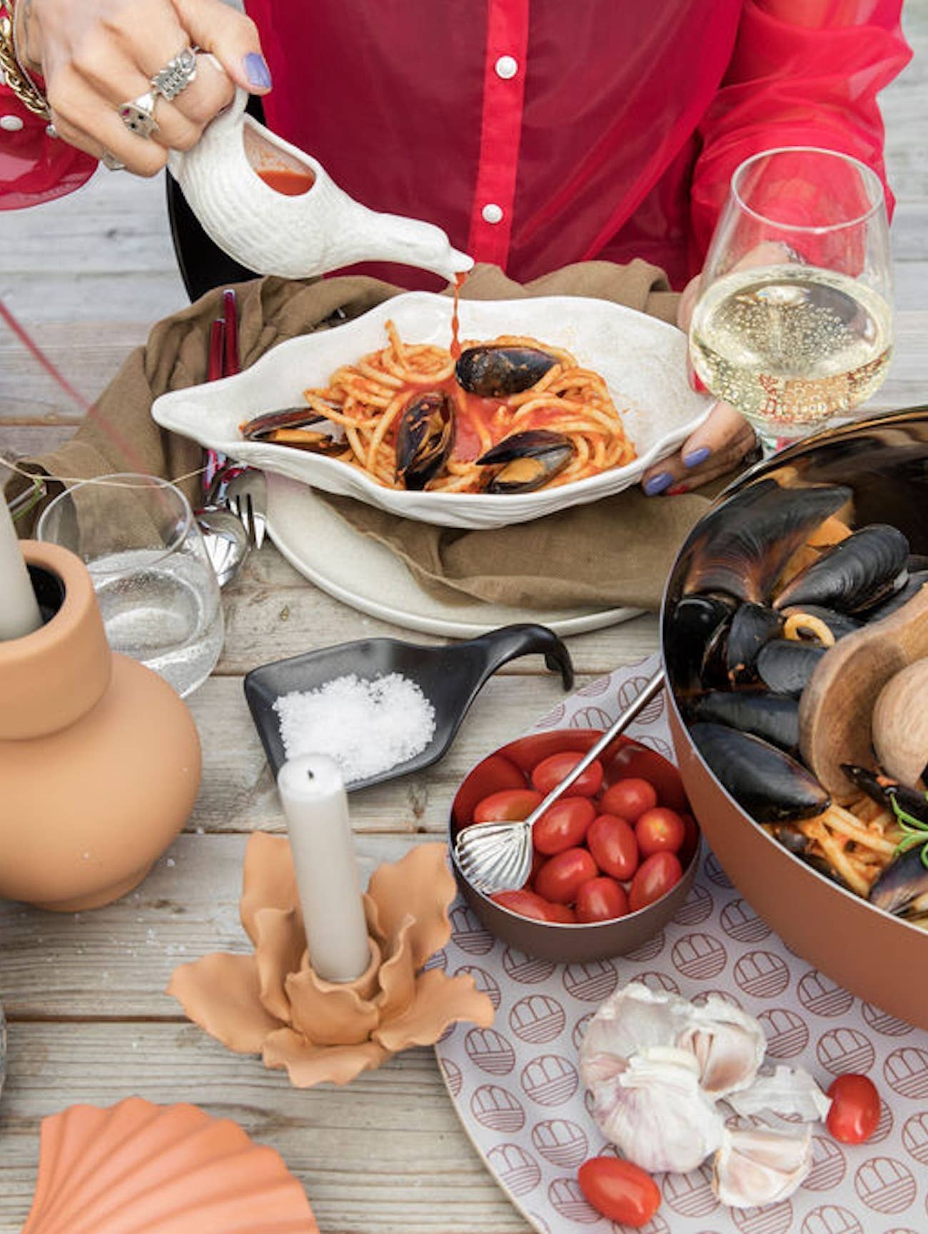 Eine Person in einem roten Hemd gießt Soße über Muschelnudeln, die auf einer BYON Design Schale Alima von Sagaform GmbH serviert werden, umgeben von Weißwein, Kirschtomaten, Knoblauch, Salz, Wasser und einem Topf mit weiteren Muschelnudeln auf einem Holztisch.