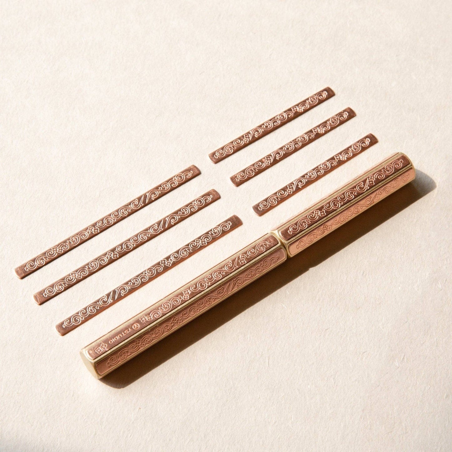 Zwei ENTREPÔT - YSTUDIO Klassische Renaissance KAZARI KANAGU ORCHID Tintenroller mit kunstvollen floralen Motiven liegen parallel auf einer beigefarbenen, strukturierten Oberfläche, begleitet von sechs koordinierten Stiftkappen, die alle ordentlich angeordnet sind.