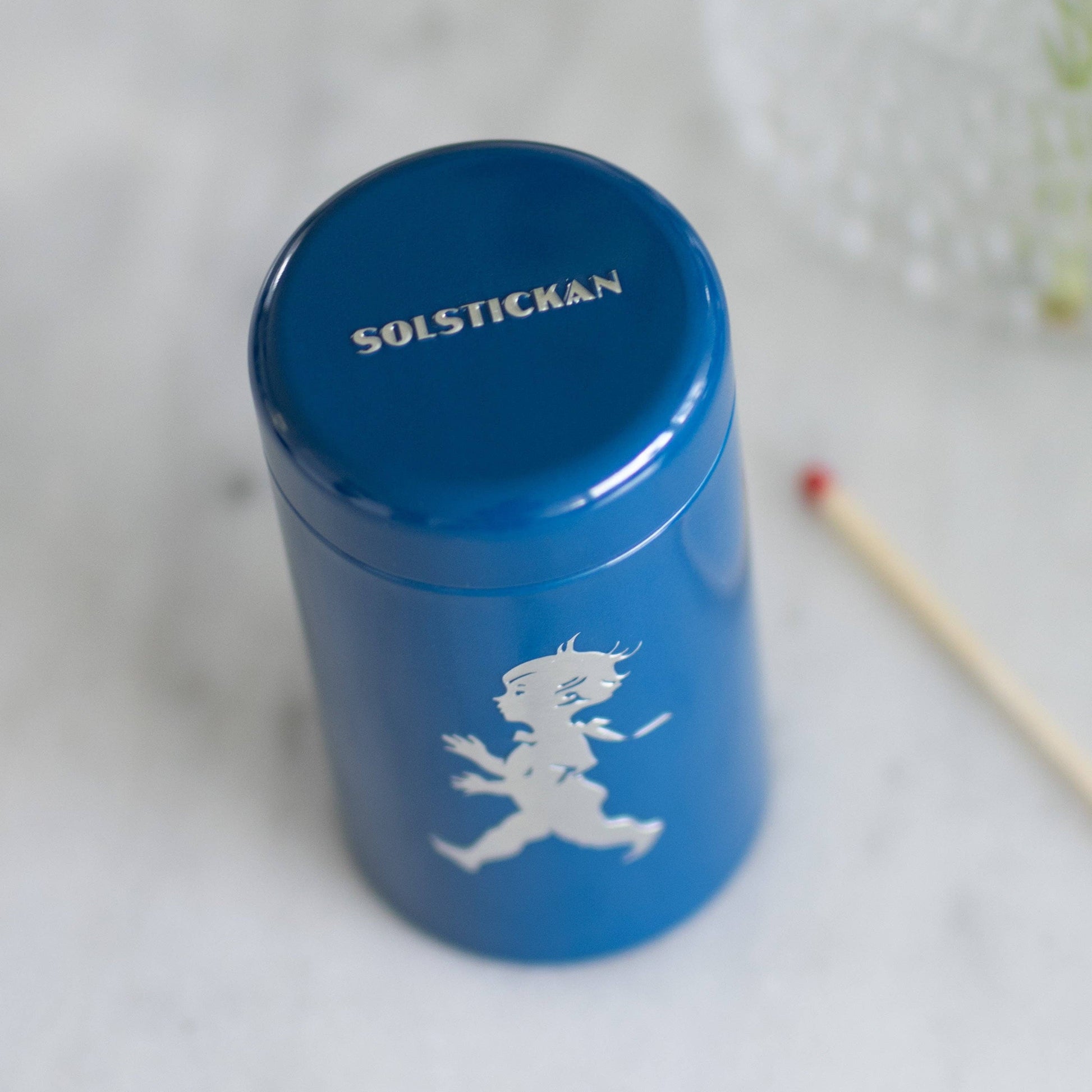 Der Solstickan Design - Passender Schlauch - Blau (nur EU) von Solstickan Design ist mit SOLSTICKAN auf dem Deckel und der kultigen Solsticke Boy-Silhouette versehen, die für echte schwedische Qualität steht. Er wird neben Streichhölzern auf einer weißen Fläche präsentiert.