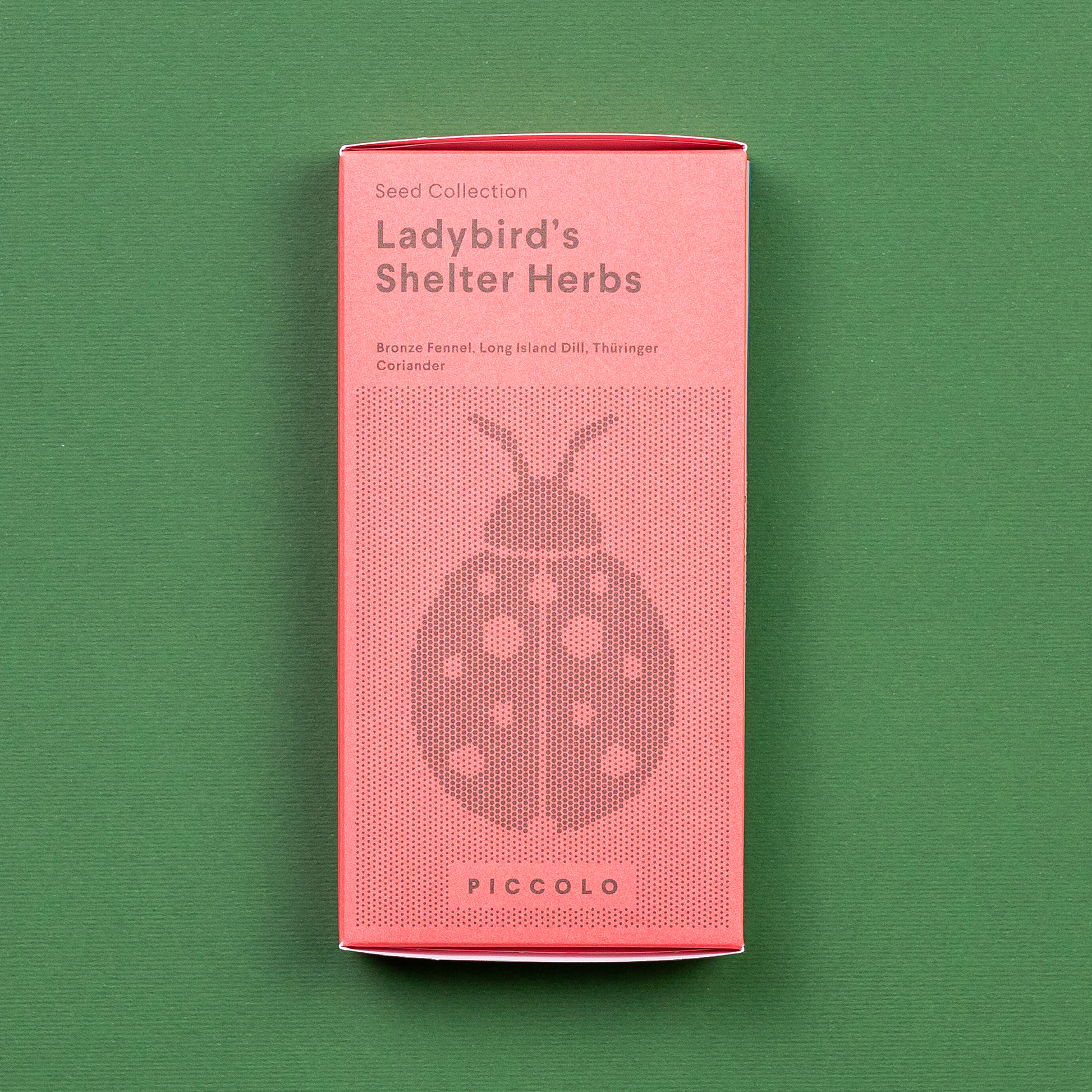 Eine rosafarbene Piccolo Seeds - Ladybird's Shelter Kräuter-Packung mit einer Marienkäfer-Illustration ruht auf Grün. Es hebt Samen hervor: Fenchel, Dill und Thaiingher Koriander der Marke Piccolo Seeds.