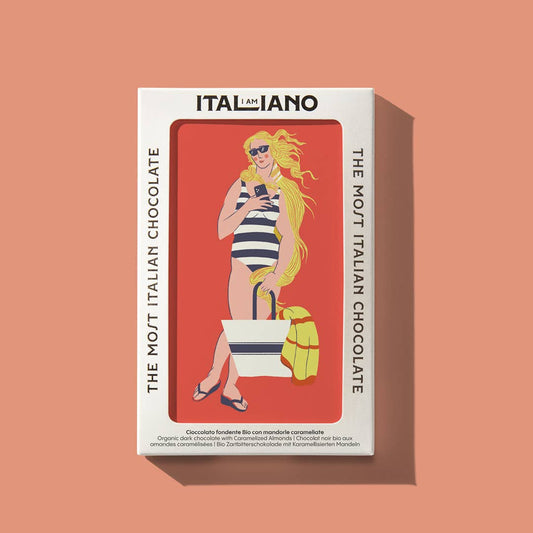 Eine Schachtel I AM ITALIANO - Venere Bio Zartbitterschokolade mit Mandel zeigt eine Frau im gestreiften Badeanzug, die eine Tasche und ein Handtuch auf einem korallenroten Hintergrund hält. Text: "ITALIANO" und "THE MOST ITALIAN CHOCOLATE". Feine Bio-Schokolade, karamellisierte Mandel.
