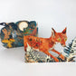 Die Fuchs Collage Karte von Mark Hearld by Art Angels zeigt einen roten Fuchs mit gelben Augen inmitten von Farnen und einen weiteren Fuchs unter einem Vollmond in einem sternenklaren Nachthimmel, der Hearlds unverwechselbaren Collage-Stil einfängt.