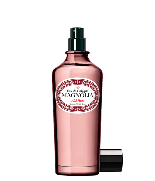 Ein rosafarbener Glasflakon von Ach.Brito Eau de Cologne Magnolia 100 ml mit dunklem Sprühkopf, weißem Etikett mit roter und schwarzer Schrift und daneben liegendem Verschluss, der den Duft andeutet.
