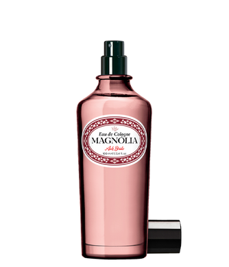 Ein rosafarbener Glasflakon von Ach.Brito Eau de Cologne Magnolia 100 ml mit dunklem Sprühkopf, weißem Etikett mit roter und schwarzer Schrift und daneben liegendem Verschluss, der den Duft andeutet.