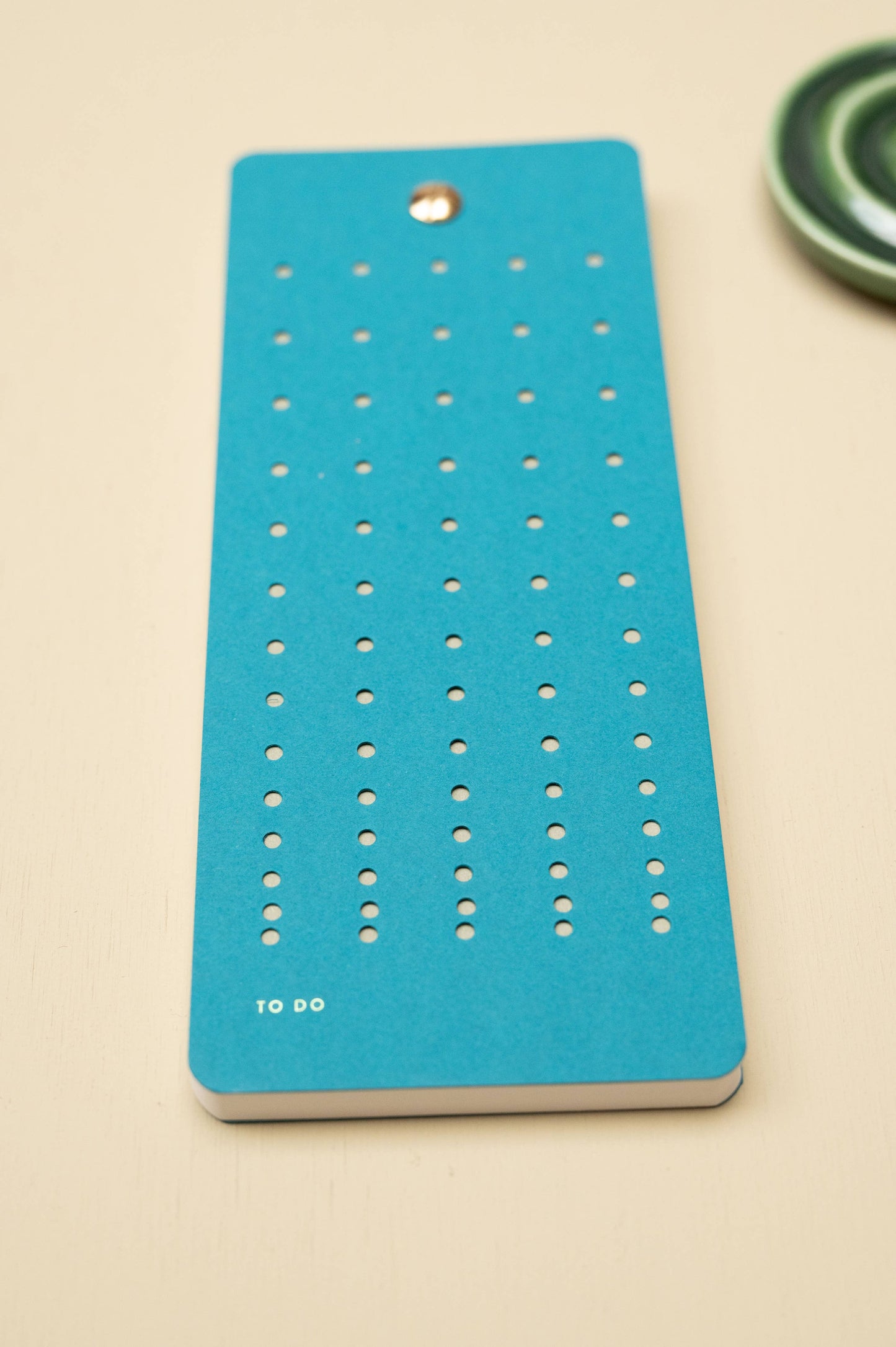 Die Tinne Mia - Aufgabenliste - Blaugrün von Tinne Mia ist ein blau-grüner, rechteckiger Notizblock aus FSC®-zertifiziertem Papier mit kreisrunden Löchern und dem Schriftzug TO DO" in weißer Schrift unten links auf einer hellen Oberfläche.