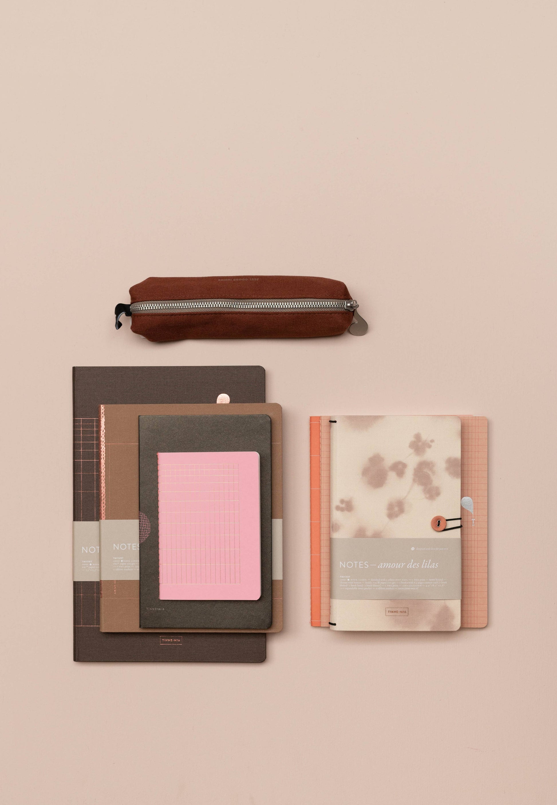 Ein braunes Mäppchen mit Reißverschluss sitzt auf vier Tinne Mia Notizbuchknopf Leinen "Amour des lilas" FSC Mix Planern und Bullet Journals in Braun, Rosa und Beige, ordentlich angeordnet auf einem hellbeigen Hintergrund.