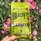 Eine Hand hält eine leuchtend grüne Tüte Brady's Coffee Company - Brady's Celtic Blend Kaffee vor rosa Blumen. Das Etikett trägt die Aufschrift „Irish Roast“, „Co. Wicklow“ und hebt die Spezialitätenqualität hervor.