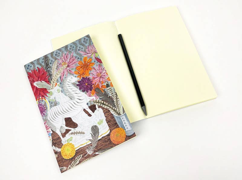 An Art Angels Notizbuch mit Zebra-Dahlien und Federn von Angie Lewin liegt teils auf einem offenen, unformatierten Notizbuch; ein schwarzer Stift ruht auf den Seiten, alles arrangiert auf weißem Untergrund.