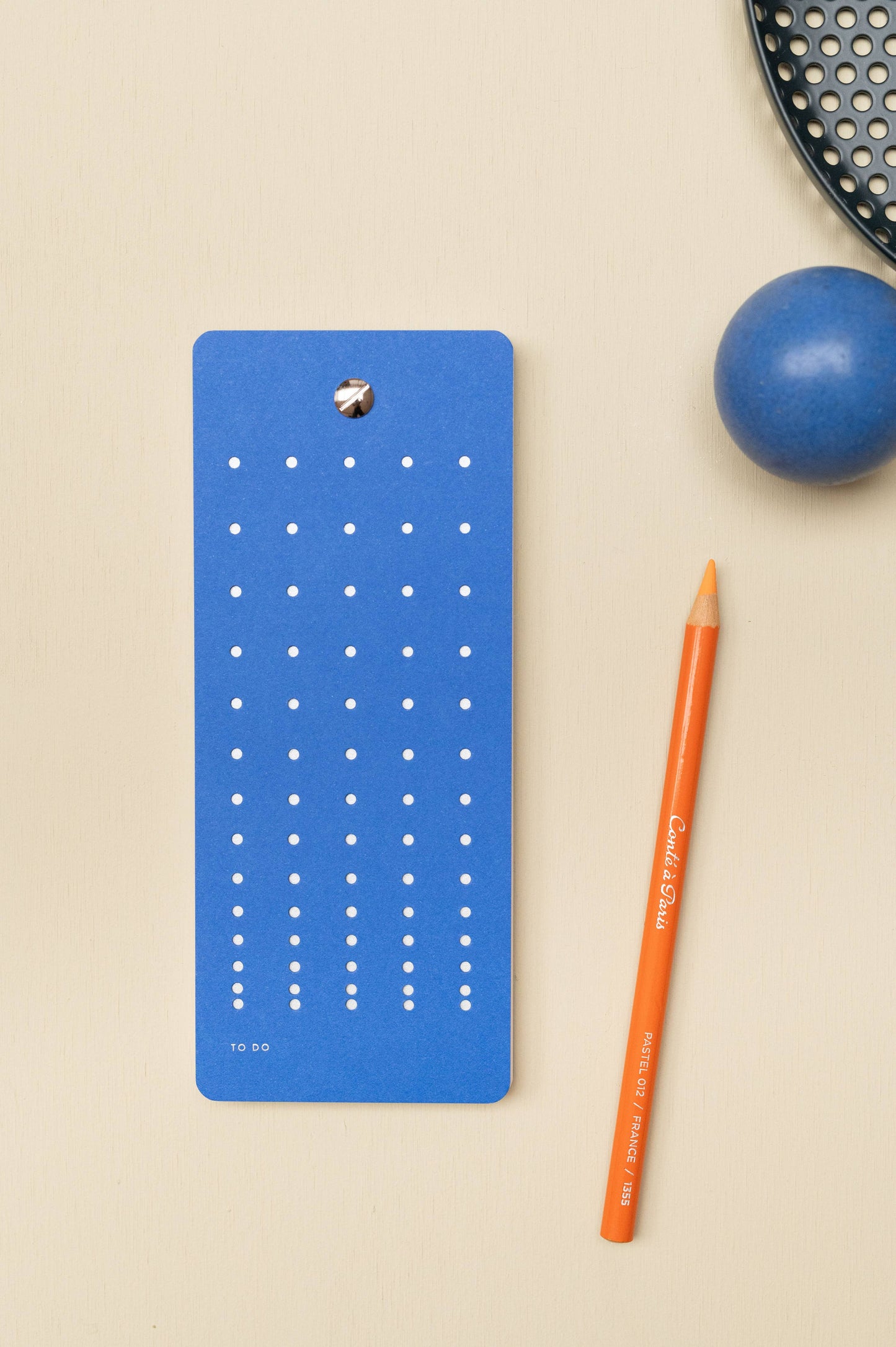 Die Tinne Mia - Aufgabenliste - True Blue von Tinne Mia, hergestellt aus FSC-Papier, ruht auf einer beigen Fläche neben einem orangefarbenen Bleistift. In der rechten oberen Ecke erscheinen eine blaue Kugel und ein Teil eines schwarzen, runden Tabletts.