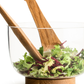 In einer gläsernen Salatschüssel mit Holzboden, gefüllt mit Blattgemüse, ruht das Sagaform GmbH Nature Salatbesteck, kurz, 2-teilig.