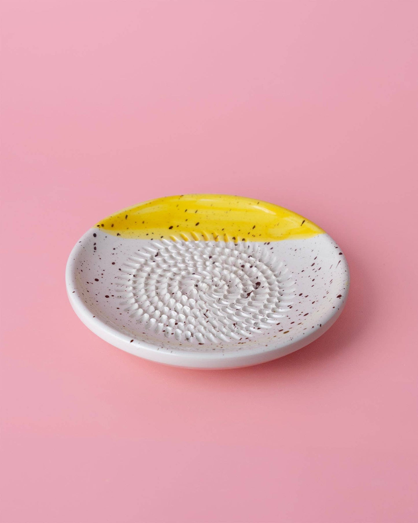 Der Alfar Tierra Cocida | Kleiner Keramikreibe Teller ZIMT ist ein kleiner, runder, handgefertigter Keramikteller mit weiß gesprenkelter Oberfläche, spiralförmiger Textur und gelbem Akzent - ideal als Weihnachtsgeschenk. Abgebildet auf einem rosa Hintergrund.
