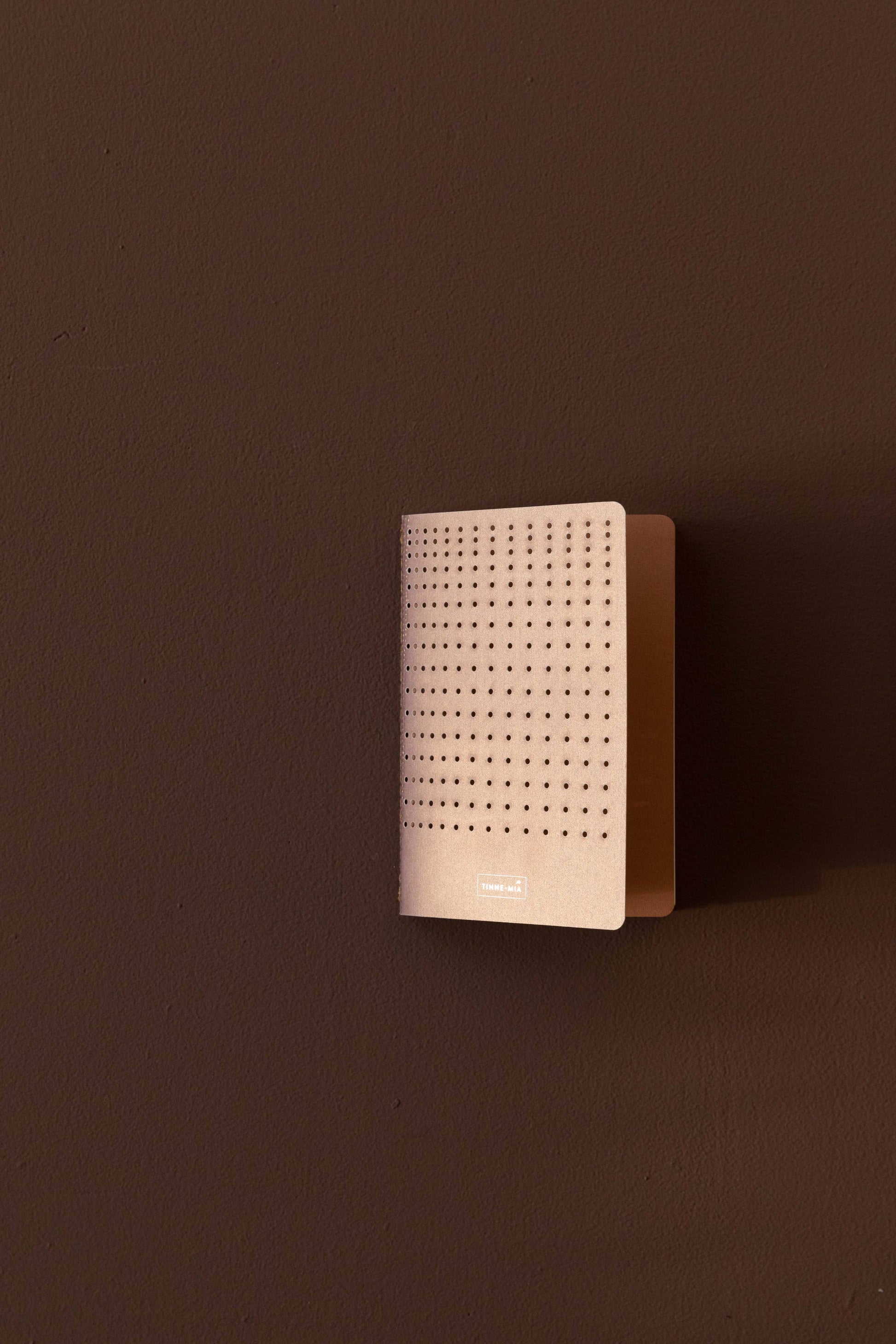 Ein kompaktes, rechteckiges Tinne Mia - Bullet Journal A6 in Grid Burgunderrot (FSC Mix) mit minimalem Design ruht auf einer braunen Wand, die einen dezenten Schatten und ein Etikett im unteren Bereich aufweist.