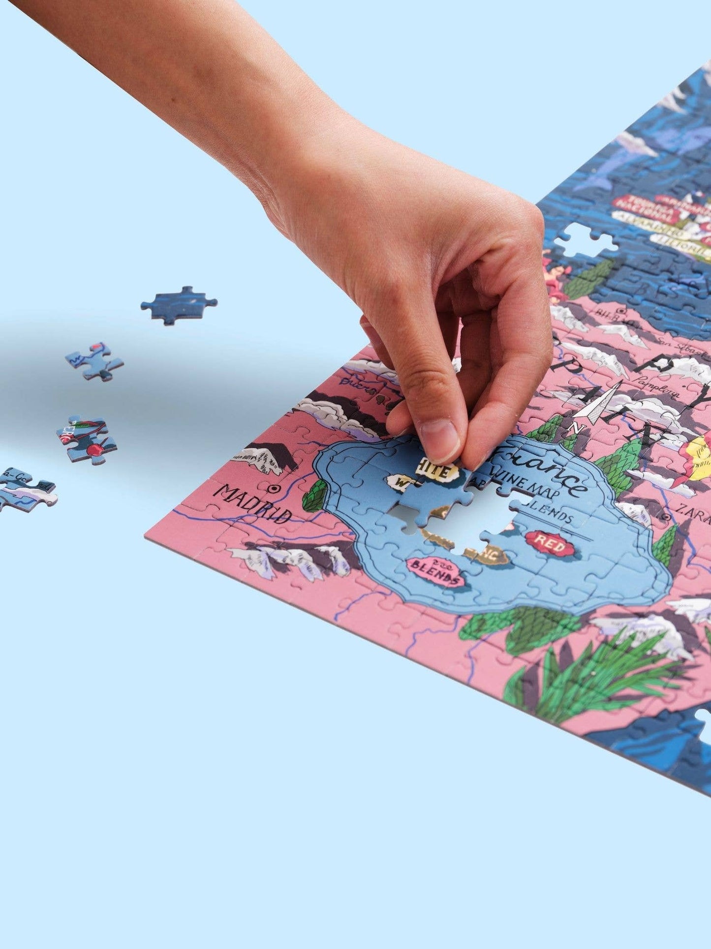 Eine Hand legt ein Teil in das bunte Water & Wines - Wein Puzzle - Frankreich von Water & Wines, das eine Karte mit Bezeichnungen wie Madrid und französische Weinregionen zeigt, während lose Teile auf einem hellblauen Hintergrund liegen.