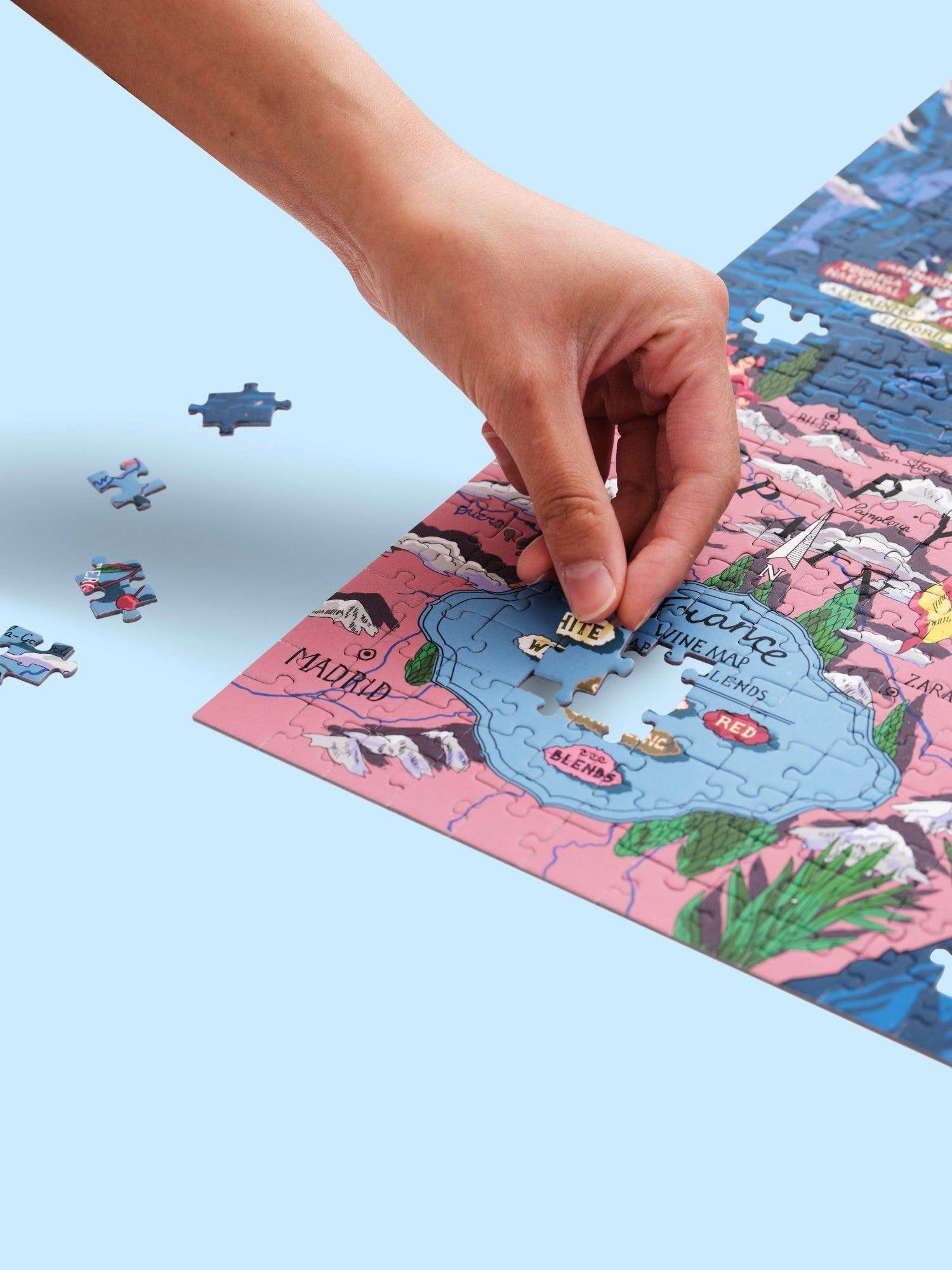 Eine Hand legt ein Teil in das bunte Water & Wines - Wein Puzzle - Frankreich von Water & Wines, das eine Karte mit Bezeichnungen wie Madrid und französische Weinregionen zeigt, während lose Teile auf einem hellblauen Hintergrund liegen.