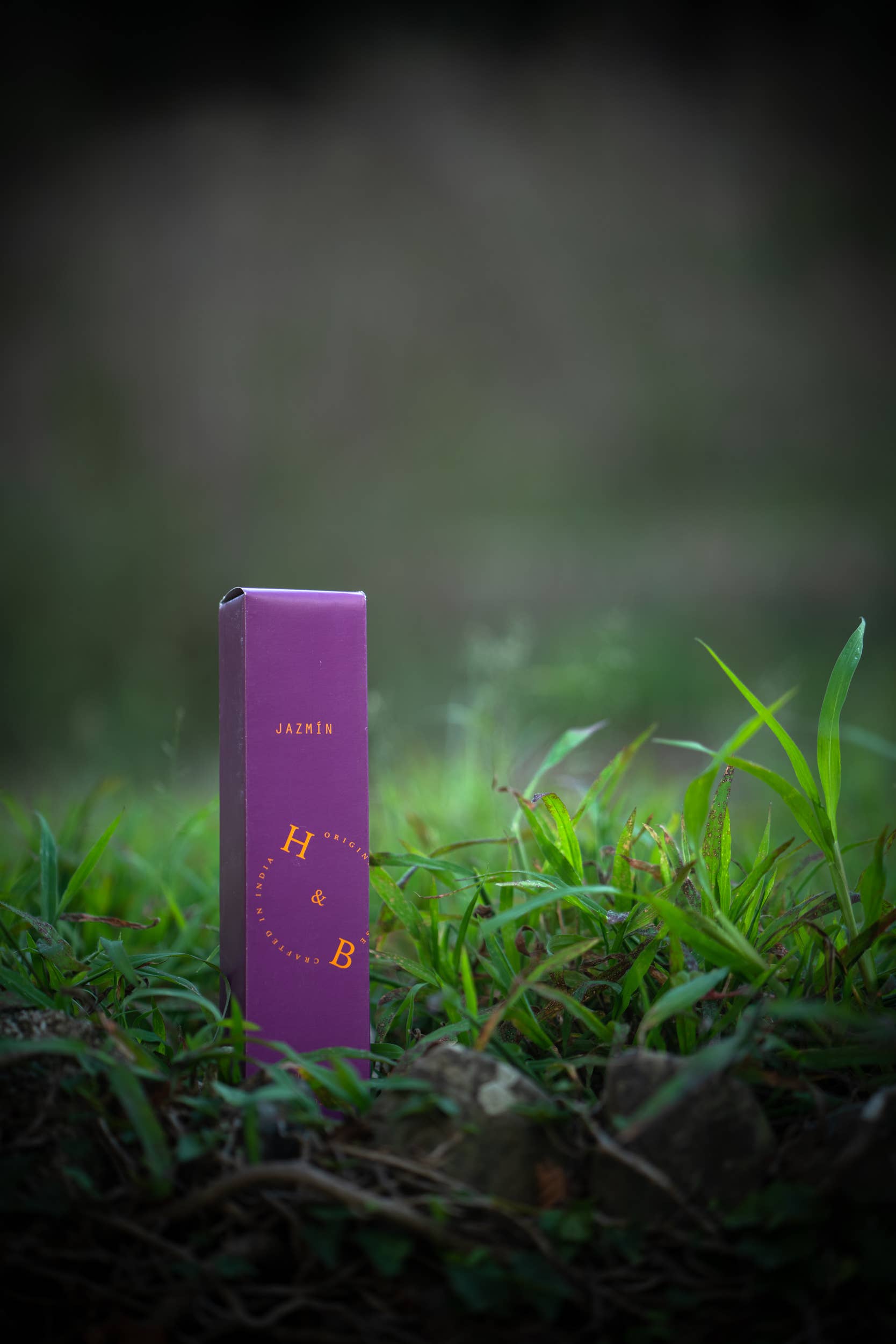 Eine lilafarbene, rechteckige H&B-Weihrauchdose mit der Aufschrift "Natürlicher Jasmin-Weihrauch" steht aufrecht auf grünem Gras mit einem unscharfen natürlichen Hintergrund, der die Essenz von aromatischen Pflanzen und reinem Jasmin einfängt.