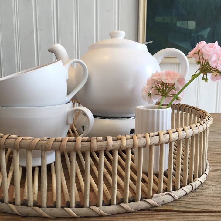 Ein Korbtablett enthält eine weiße PLINT A/S - Teekanne 1,5 Liter, zwei gestapelte weiße Teetassen und eine kleine Vase mit rosa Blumen, die alle auf einem Holztisch mit einer weiß getäfelten Wand und gerahmter Kunst im Hintergrund angeordnet sind.