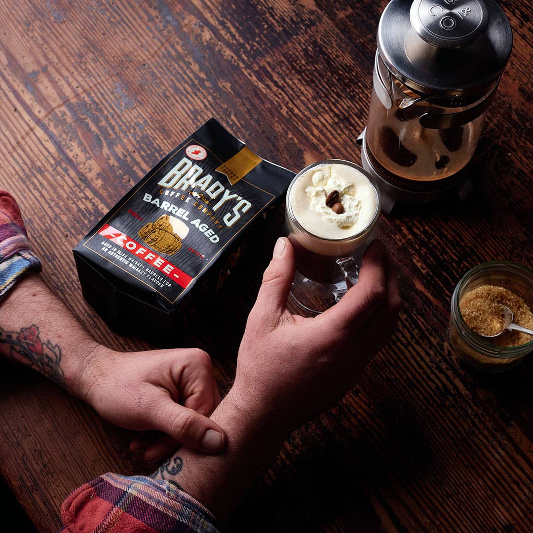 Eine tätowierte Person hält ein Glas mit cremigem Kaffee in der Hand. Auf dem Holztisch steht eine Schachtel Brady's Coffee Company - Brady's Barrel Aged Irish Whiskey Kaffeebeutel mit ganzen Bohnen, eine französische Presse, ganze Bohnen, ein Glas mit braunem Zucker und ein Löffel.