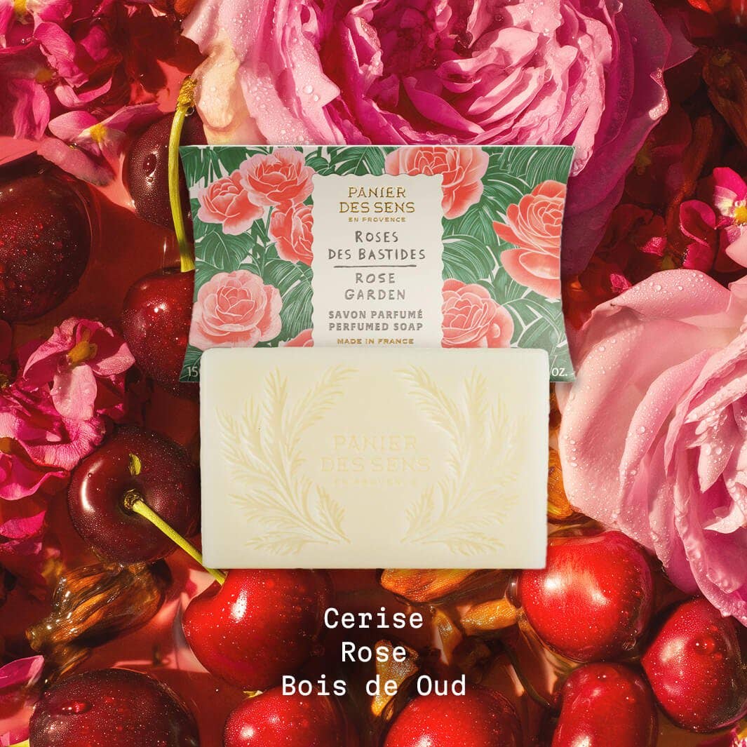 Ein Barren Panier Des Sens Duftseife | Rose des Bastides 150 g liegt vor seiner blumigen Schachtel, umgeben von frischen Rosen und Kirschen. Der Text auf dem Boden lautet: Cerise, Rose, Bois de Oud.