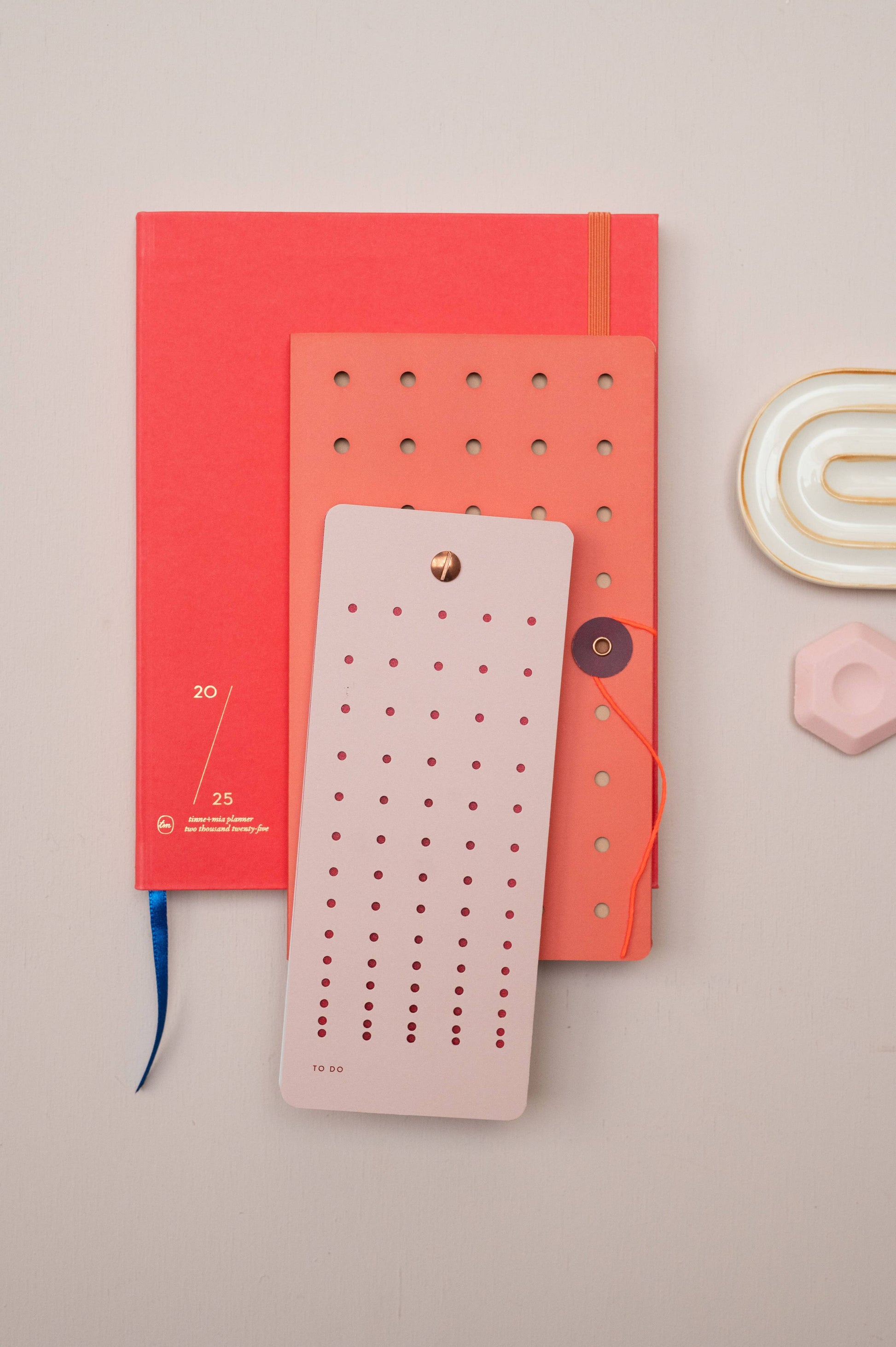 Auf hellem Untergrund liegt die Tinne Mia Aufgabenliste "Mellow Rose" mit Lochung. Daneben: ein rotes Notizbuch mit blauem Band, ein Hörbuch in Koralle (beide aus FSC-Papier), eine goldgeränderte Schale und ein pinkes Sechseck.
