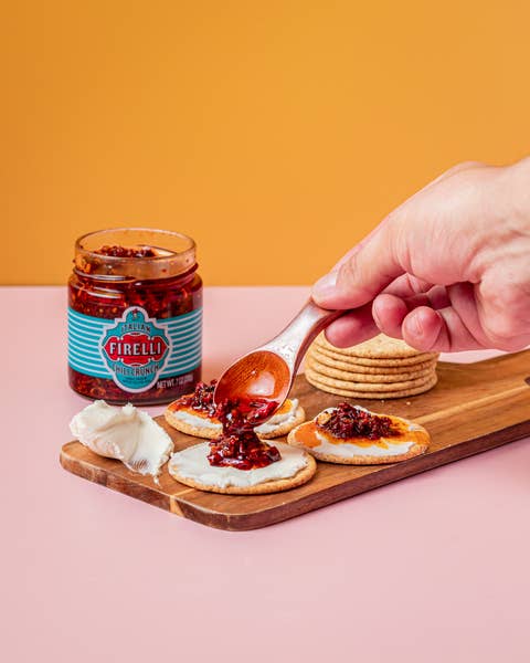 Eine Hand verteilt Firelli - Casa Firelli Chili Crunch 200g Glas mit einem Löffel auf einem Frischkäsecracker. Der Cracker liegt auf einem Holzbrett mit weiteren Crackern, Frischkäse und einem Glas Firelli Chili Crunch auf einer rosa Fläche vor orangefarbenem Hintergrund.