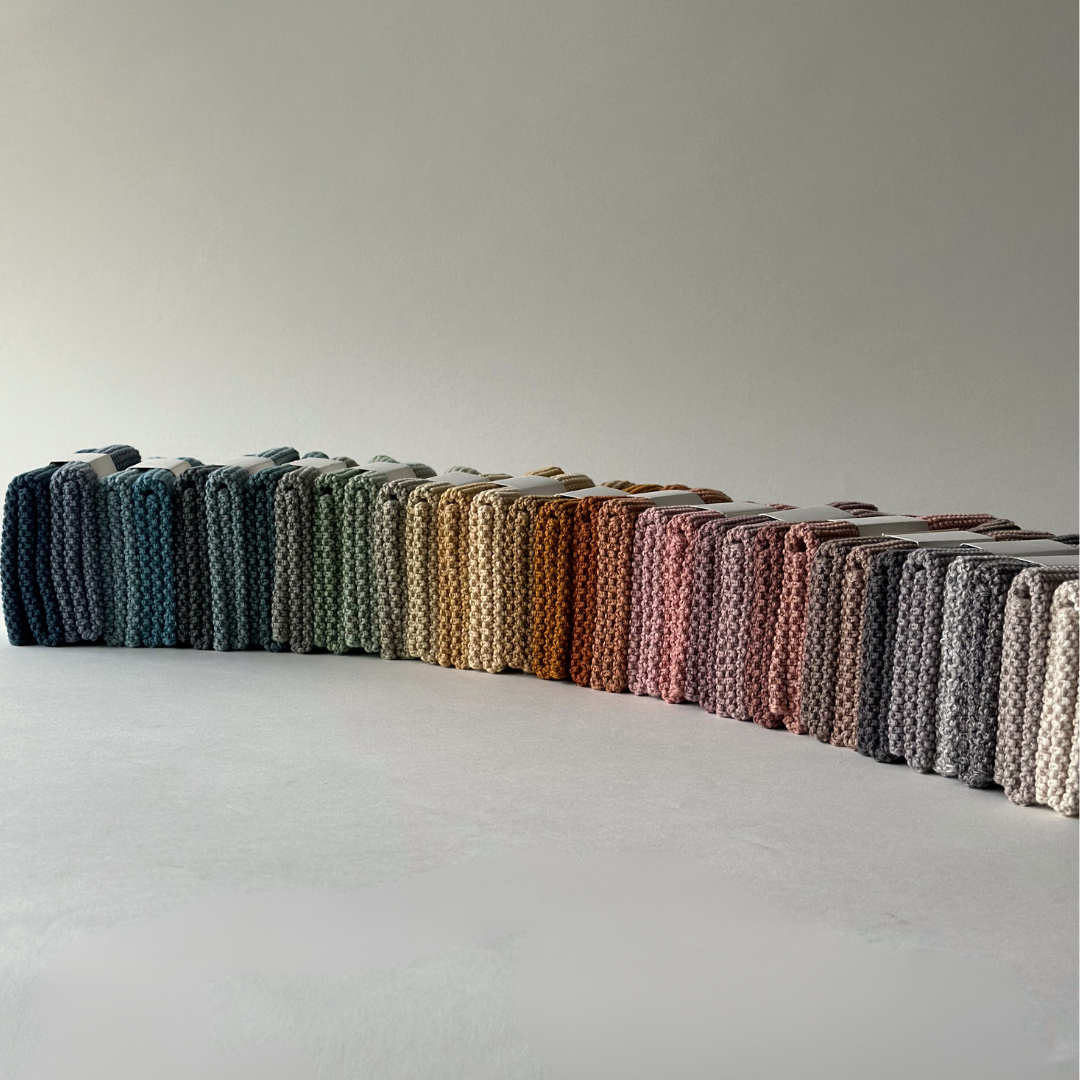 Reihen von ordentlich gefalteten Aspegren Denmark - Spültücher, gestrickt, massiv, Kiefernholz in verschiedenen Farben - dunkelblau und grün auf der linken Seite bis rosa, hellbraun und grau auf der rechten Seite - bilden ein umweltfreundliches Display vor einem schlichten Hintergrund.