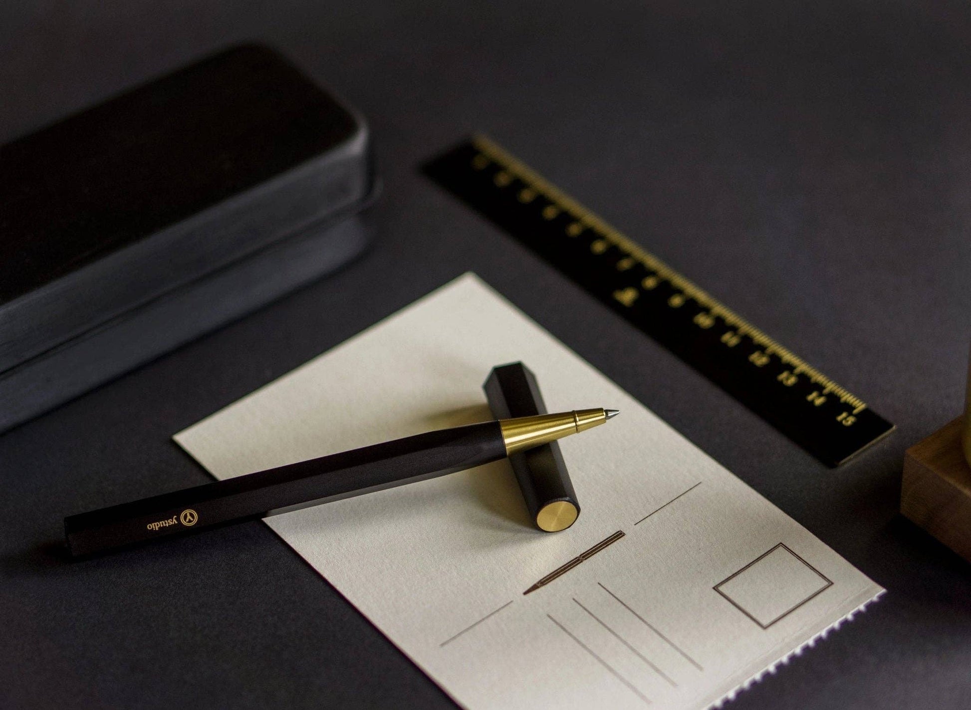 Der ENTREPÔT - YSTUDIO Harz-Rollerball-Stift (Schwarz) ruht auf einer gefütterten Notizkarte, die Kappe liegt daneben, dazu gesellen sich ein schwarzes Etui und ein schwarzes Lineal mit goldenen Markierungen, so dass ein elegantes Schreibritual auf dunkler Fläche entsteht.