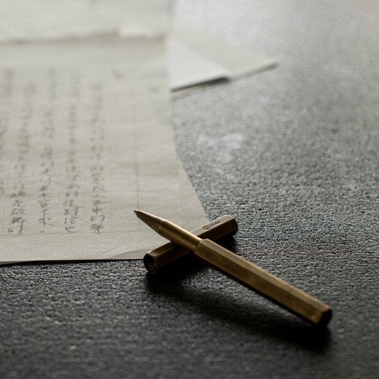 Der ENTREPÔT - YSTUDIO Klassischer Revolve-Rollerball-Stift aus Messing von ENTREPÔT liegt auf einer dunklen, texturierten Fläche bei natürlichem Licht neben Papieren mit asiatischen Schriftzeichen.