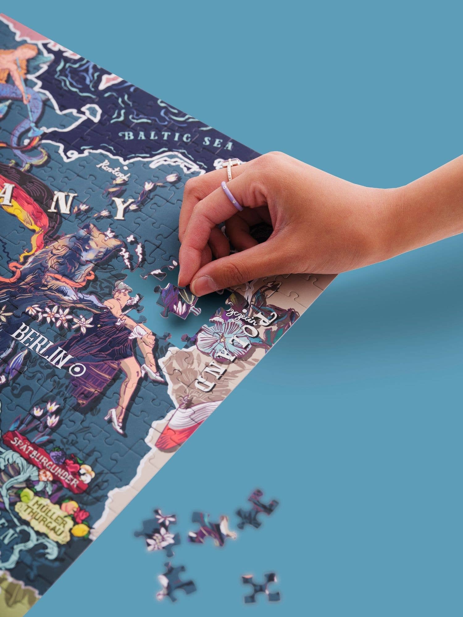 Eine Hand legt ein Teil in ein teilweise fertiges Water & Wines - Wein Puzzle - Deutschland auf einer blauen Fläche, mit losen Teilen in der Nähe - eine spannende Art, deutsche Weinregionen zu entdecken.