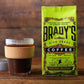 Ein Glas Kaffee mit Korkhülle steht neben einer hellgrünen Tüte Brady's Coffee Company – Brady's Celtic Blend Kaffee, einer irischen Röstung von Brady's Coffee Company, die auf einer Holzoberfläche wahre Spezialitätenqualität verspricht.