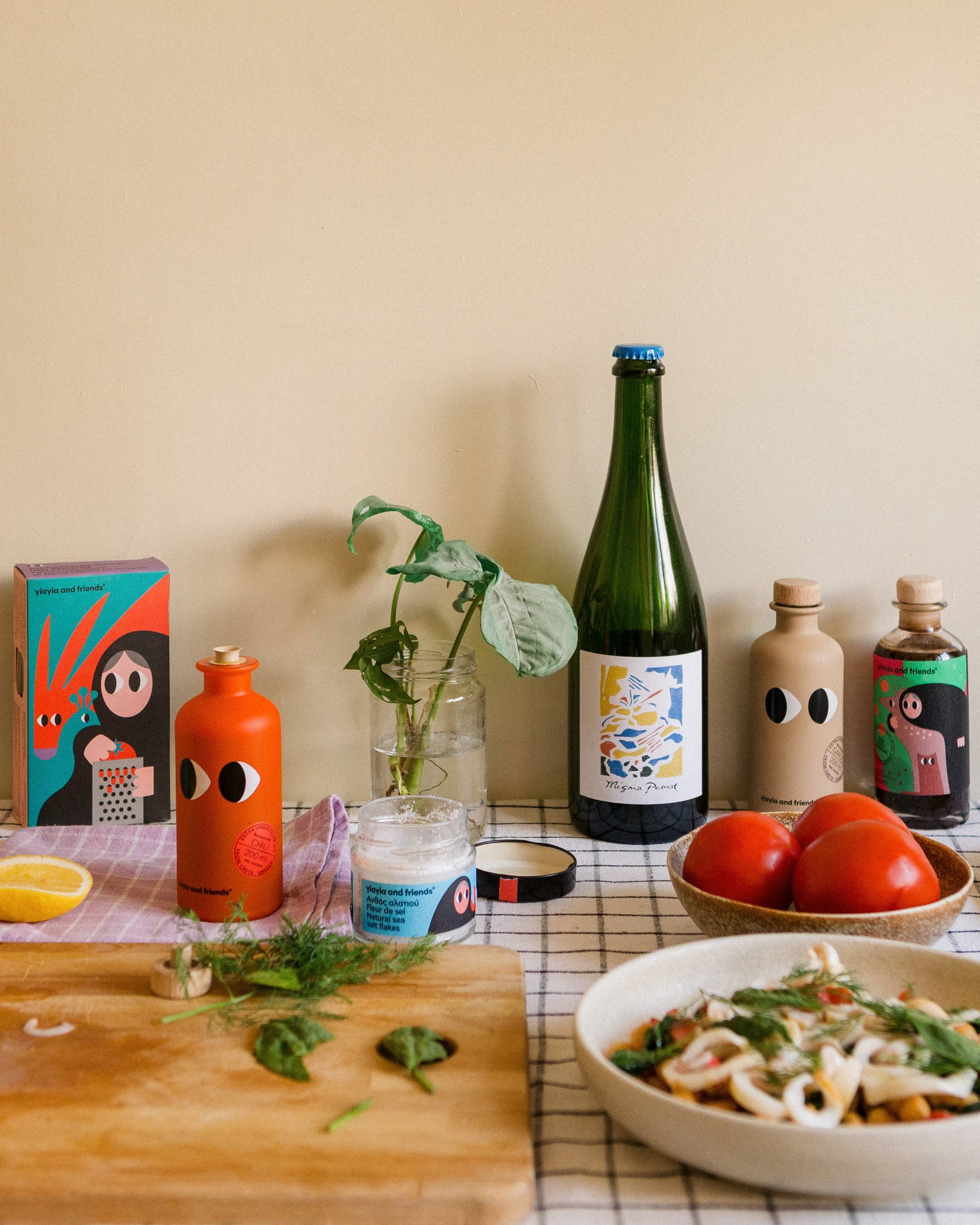 Eine Küchentheke mit Flaschen von yiayia and friends - Natives Olivenöl extra mit Chili, ein Glas mit Kräutern, Tomaten, ein Schneidebrett mit Kräutern und Zitrone, ein bunter Kunstkasten, eine Schüssel mit Nudeln und ein kariertes Handtuch vor einer beigen Wand.