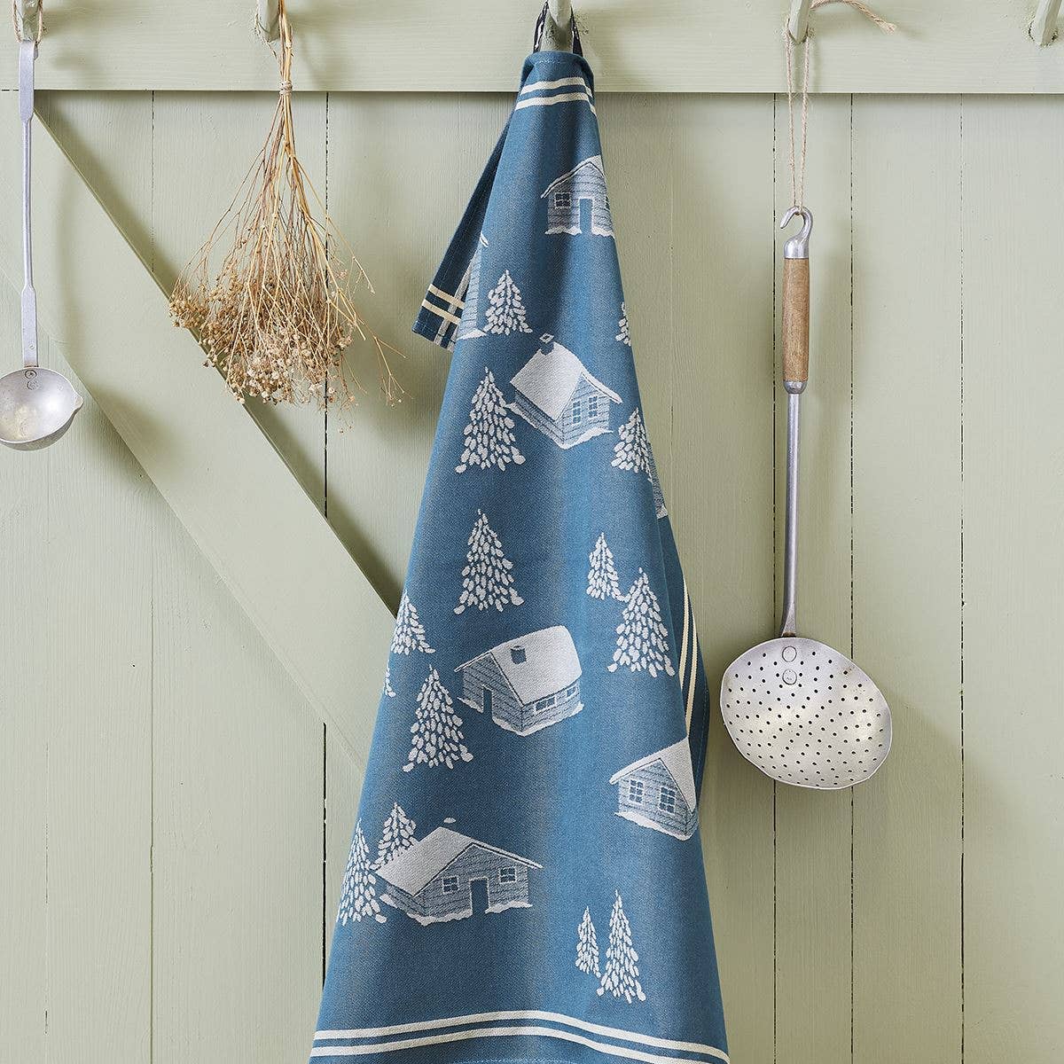 Das Geschirrtuch Sylvie Thiriez - Torchon Jacquard Coeur Vallée Blau aus OEKO-TEX-zertifizierter Baumwolle mit weißen Hütten- und Baummotiven hängt an einer hellgrünen Wand neben Küchenutensilien und getrockneten Kräutern.