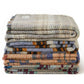 Ein Stapel von vier Tweedmill Textiles Zufällige recycelte Wolldecke - Groß in verschiedenen Beige-, Grau-, Blau- und Rotmustern, hergestellt in Großbritannien, ausgestellt vor einem weißen Hintergrund.