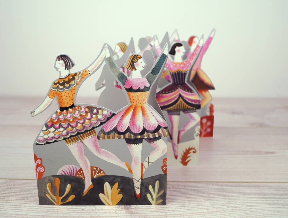 Scherenschnitte von Ballerinas in bunten Kostümen stehen auf einer hellen Holzoberfläche. Die Tänzer Ausstanzkarte von Sarah Young für Art Angels zeigt florale und abstrakte Muster und ist ein charmantes Design-Geschenk für jeden Anlass.