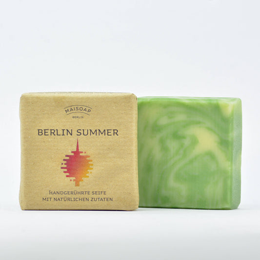 Ein Riegel grün-weiß verwirbelter maisoap berlin - Berlin Summer Seife steht neben seiner braunen Papierverpackung mit einer rot-gelben Grafik und deutschem Text der Marke maisoap berlin vor einem schlichten weißen Hintergrund.