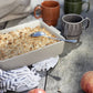 Ein rechteckiger Fruchtstreusel mit Servierlöffel steht auf einem rustikalen Tisch, umgeben von zwei Sagaform Coffee & More Kaffeebecher 2-teilig der Sagaform GmbH, einer Stoffserviette und zwei Äpfeln.