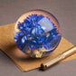 Der Briefbeschwerer mit Kornblume von Hafod Grange ist eine Glaskugel mit blauen Trockenblumen, ideal als Schreibtisch-Accessoire - hier platziert auf braunem Papier neben einem Goldstift auf grauem Untergrund.