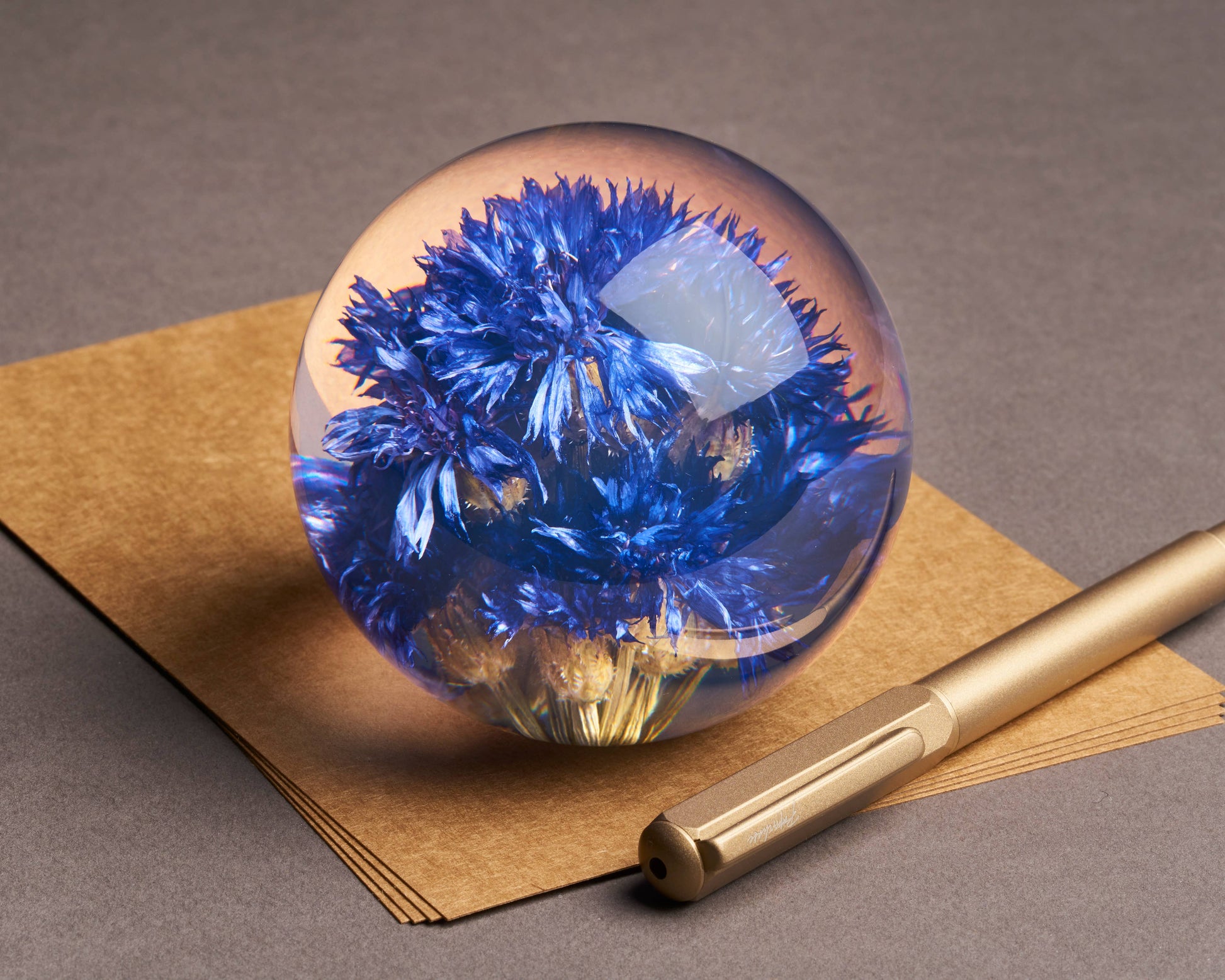 Der Briefbeschwerer mit Kornblume von Hafod Grange ist eine Glaskugel mit blauen Trockenblumen, ideal als Schreibtisch-Accessoire - hier platziert auf braunem Papier neben einem Goldstift auf grauem Untergrund.